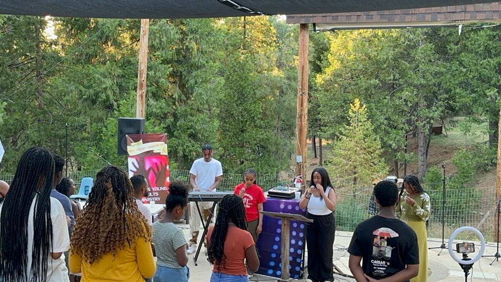 Young adults leading outdoor worship with keyboard and vocals under a canopy in the forest