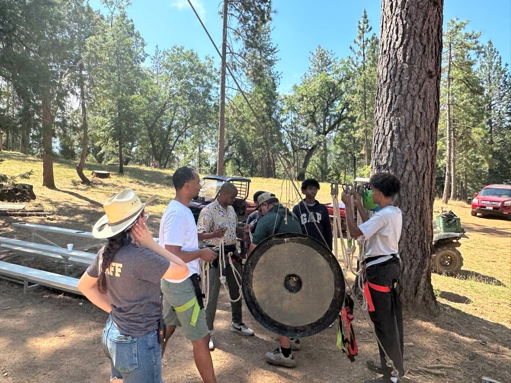 Youth and staff preparing climbing ropes and gear beside a large tree at camp