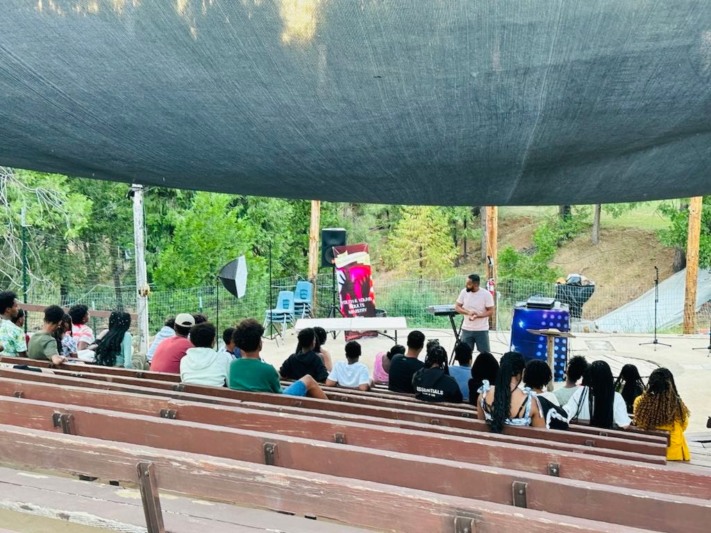Youth seated on outdoor amphitheater benches listening to a speaker at a camp session